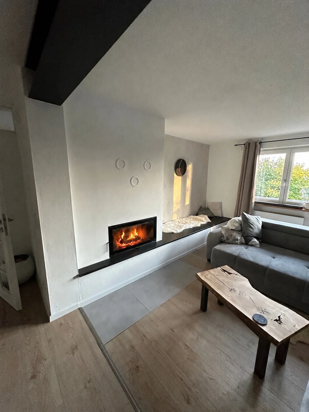 Moderner verputzter Grundofen mit großer Sichtscheibe und schwarzer Natursteinbank in Kröning. Feuer mit KI bearbeitet.